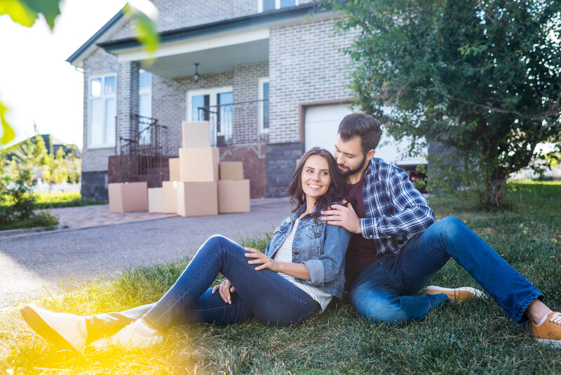 man and woman sitting on the lawn in the front of their house with moving boxes behind them